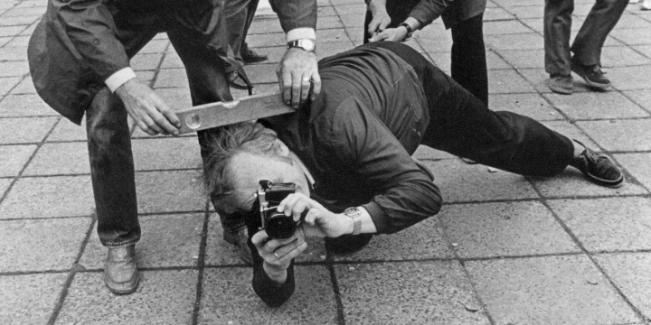 Schwarz-weiß Foto eines Mannes, der auf dem Boden liegt und etwas im Vordergrund fotografiert, während ihm ein anderer Mann eine Wasserwaage auf den Kopf legt