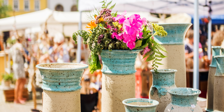 Eine Gruppe aus vier blau-braunen Vasen mit einer Blume im Vordergrund und im Hintergrund Stände auf dem Jenaer Töpfermarkt, der auf dem Marktplatz stattfindet. 