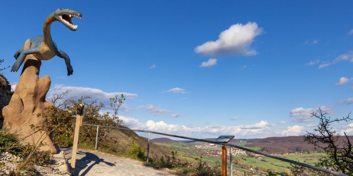 Ein Dinosaurier auf einem Felsen am Wegesrand im tal sind Häuser zu sehen. Kleine Wolken sind amblauen Himmel. 