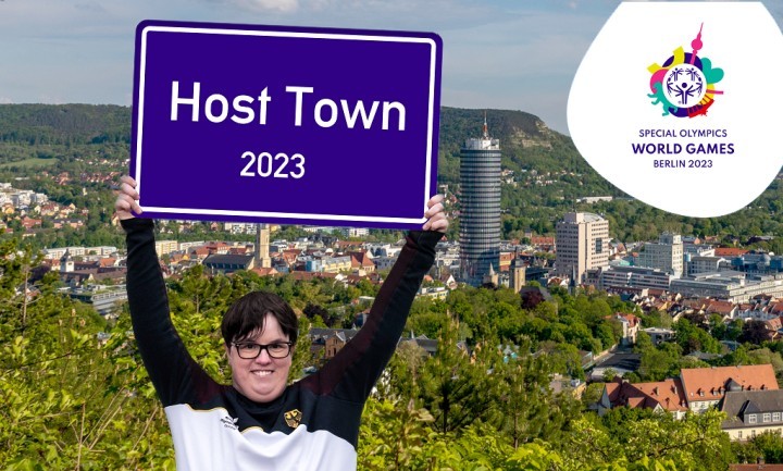 Frau in offizieller Sportkleidung mit einem Schild "Host Town 2023" vor einem Panoramafoto der Stadt Jena