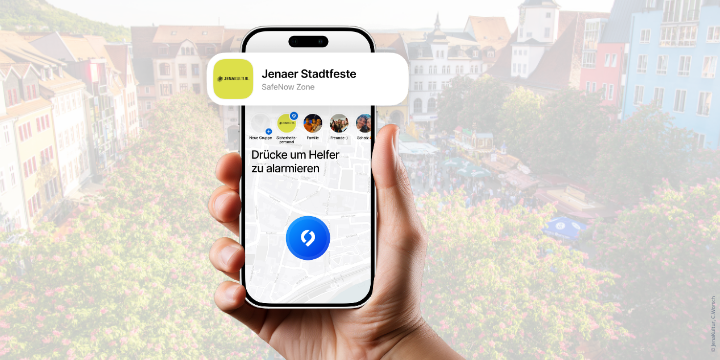 Hand mit Handy, auf desssen Bildschirm die SafeNow-Zone zu sehen ist. Im Hintergrund ist der Jenaer Markt mit Bühne und Punblikum von oben zu sehen.