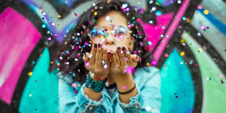 Junge Frau in Jeansjacke steht vor einer mit buntem Graffiti besprühten Wand und pustet aus ihren erhobenen Händen Konfetti in die Kamera  ©Unsplash