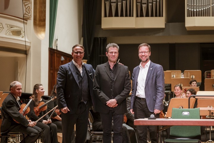 Carsten Müller, Simon Gaudenz und Thomas Nitzsche stehen vor dem Orchester der Jenaer Philharmonie neben der Unterzeichnung der Vertragsverlängerung von Simon Gaudenz