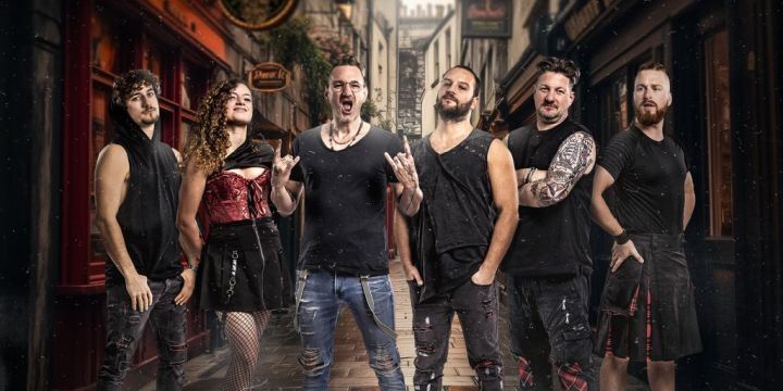 Die Band besteht aus 5 Männern und einer Frau. Sie stehen vor dem Hintergrund einer Gasse. Links und rechts sind Kneipen zu erkennen.