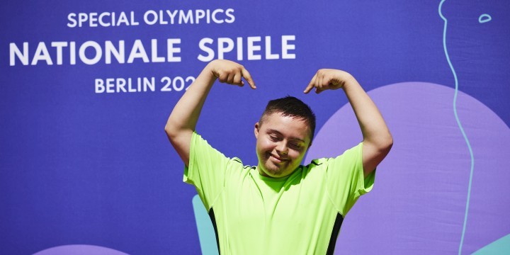 Athlet mit Trisomie 21 steht vor einer lila Pressewand mit der Aufschrift "Special Olympics Nationale Spiele Berlin 2023" und hebt beide Arme über seinen Kopf, um auf sich zu zeigen  ©Anna Spindelndreier