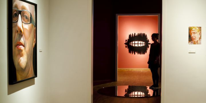 Ausstellungsbereich in der Kunstsammlung mit zeitgenössischen Gemälden und Objekten und einer Besucherin im Hintergrund