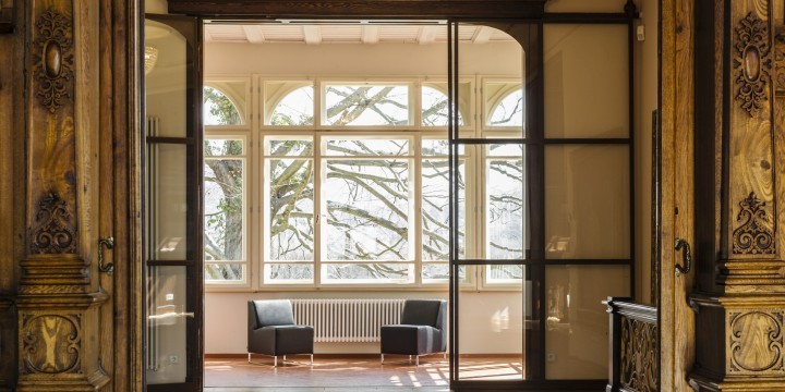 Blick durch die Flügeltür in der Villa Rosenthal auf zwei Sessel vor den Fenstern zum Garten mit Blutbuche