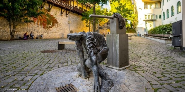 Eine abstrakter Mensch aus Bronze kniet vor einem alten Pulverturm in Jena. Auf seinem Rücken fließt durch einen danabenstehen Hahn Wasser. Dies ist der Philisterbrunner.