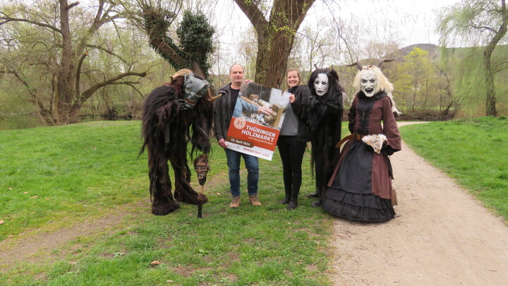 Ein Mann und eine Frau halten das Plakat des Thüringer Holzmarktes hoch und sind von mit Holzmasken als Bergteufel Menschen umrahmt.