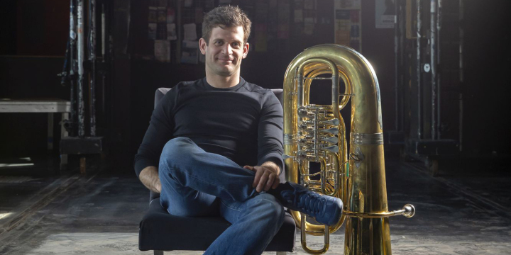 Musiker mit überschlagenem Bein sitzt auf einem Sessel auf der Bühne, die Tuba neben ihn gestellt.