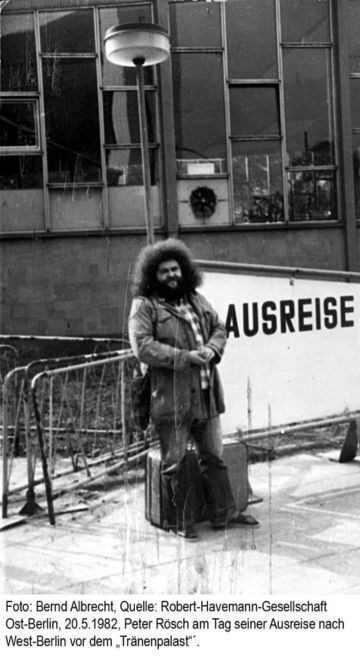 Portrait von einem Mann mit sehr lockigem Haar vor einem Gebäude mit vielen Fenstern und einem Schild mit der Aufschrift Ausreise