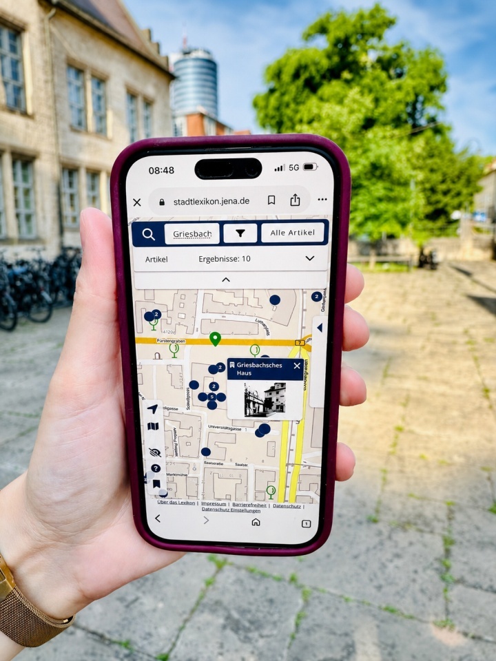 Eine Person steht in Jena an der Stelle des Griesbachschen Hauses und hat auf dem Smartphone den dazugehörigen Artikel im digitalen Stadtlexikon geöffnet