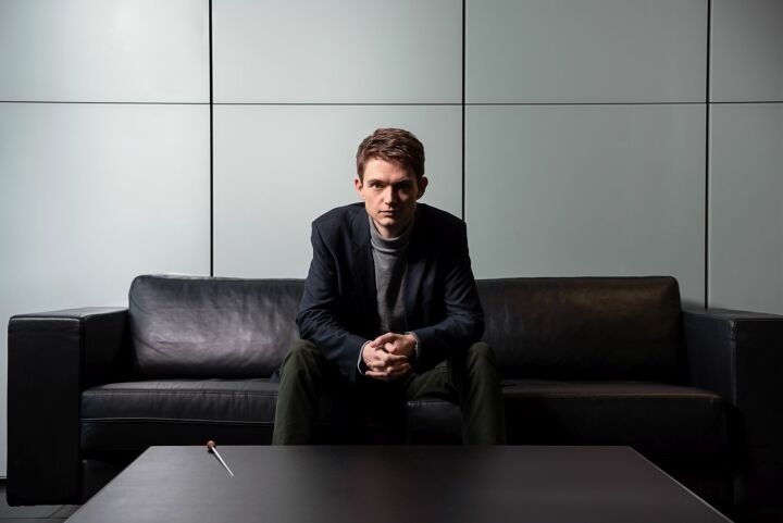 Auf Couch sitzender Musiker mit Blazer, die Hände vor seinen Schoß gefalten.