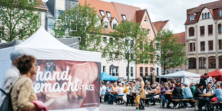 Frau und Mann laufen an einem Pavillon vorbei, auf dem handgemacht-Tage steht, im Hintergrund sitzen Gäste des Marktes.