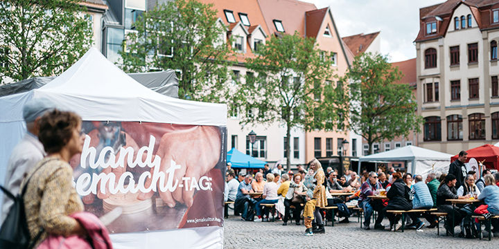 Zwei Gäste auf dem Jenaer Marktplatz laufen an einem Pavillon mit dem Aufdruck "handgemacht-Tage" entlang und weitere Gäste sitzen auf Bänken.  ©JenaKultur, C. Worsch