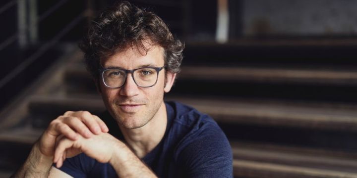 Christoph Altstaedt, Leiter des Donnerstagskonzerts sitzt auf einer Treppe. Er stützt die Ellbogen auf den Knien und hat die Hande gefaltet. Er trägt eine große Brille mit schwarzem Gestell. 