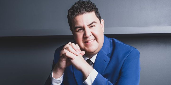 Der Musiker Tito Muñoz in einem blauen Jacket mit schwarzer Krawatte.Der Hintergrund ist in Grau gehalten. 