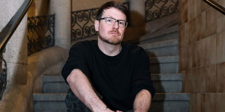 Der Musiker Robert Seidel sitzt auf der Treppe am Haupteingang des Jenaer Volksbades. Er trägt einen schwarzen Pullover und eine schwarze Jeans sowie eine Brille mit schwarzem Gestell.