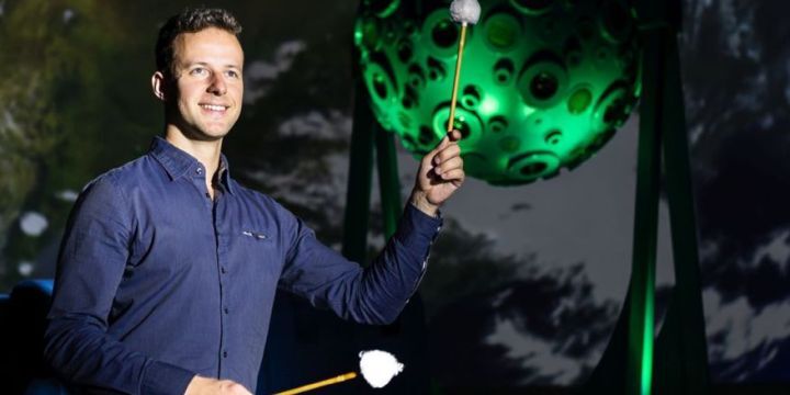Alexander Schuchert in einem blauen Hemd. Er speilt die Pauke. Hinter ihm zu sehen ist der große Projektor des Planetariums in Jena. Er wird grün angestrahlt. 