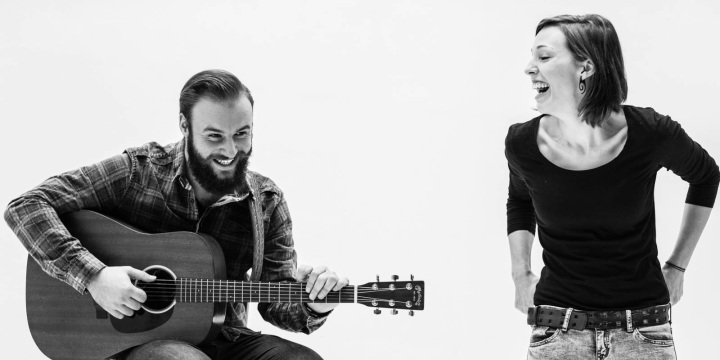 Ein Mann im Karohemd spielt sitzend Gitarre und eine Frau steht mit den Händen in den Rücken gestemmt daneben.