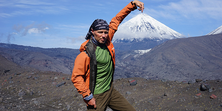 Ein Mann in Outdoorklamotten steht vor einem Berg mit schneebedeckter Kuppe und zeigt auf die Kuppe. 