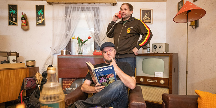 Zwei Männer stehen in einer Stube, die mit DDR-Interier ausgestattet ist; einer telefoniert und der andere liest ein Buch.