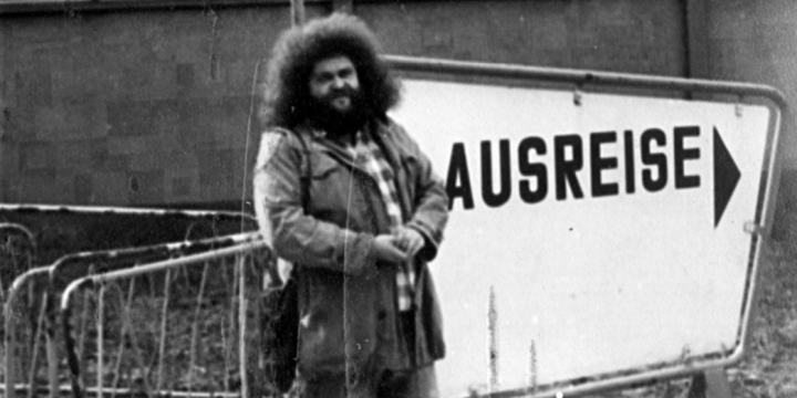 Schwarz-weiß Foto von Peter Rösch, der fröhlich an einem Zaun vor einem Schild mit der Aufschrift "Ausreise" lehnt