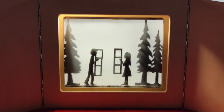 Das Foto zeigt eine Schattentheaterbühne, auf der zwei Figuren - ein junger Mann und eine junge Frau jeweils an einem Fenster stehen und sich anblicken. Im Hintergrund sind Bäume zu erkennen. 