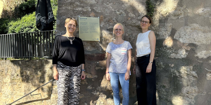 Drei Frauen an einer goldenen Tafel mit vlauer Schrift, die an einer Mauer hängt  &copy;© JenaKultur