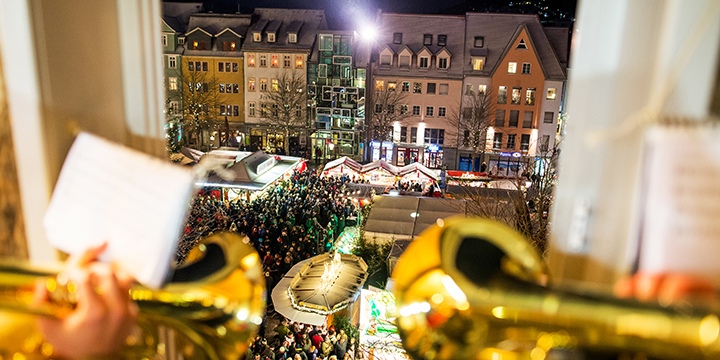 Zwei Tompeten spielen an einem Fenster, das den Blick auf den beleuchteten Jenaer Weihnachtsmarkt freigibt.